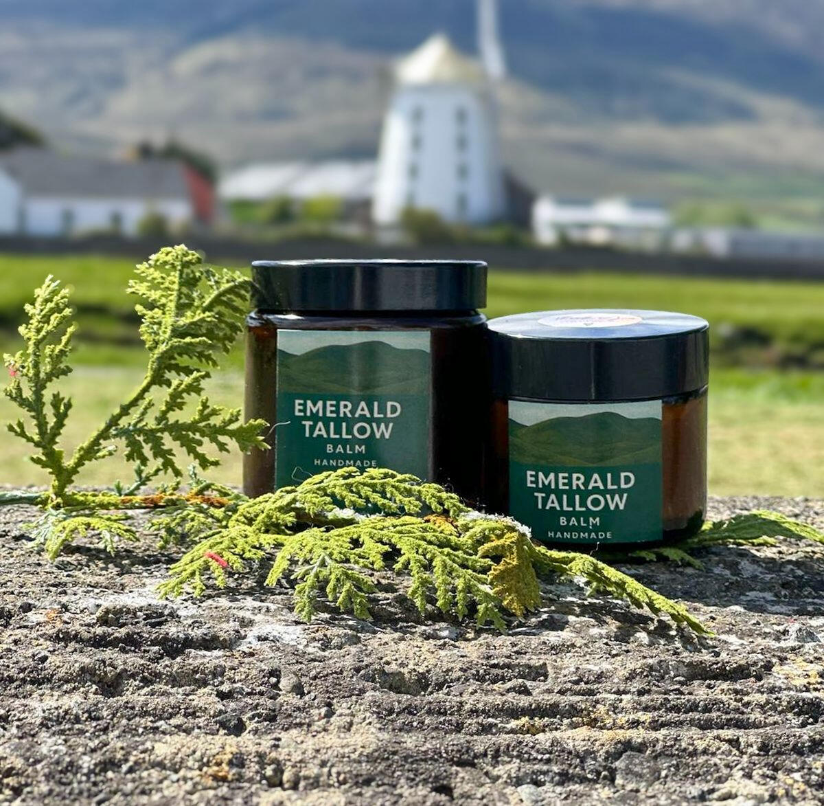 Emerald Tallow Balm Two jars of Emerald Tallow Balm on a stone wall with green foliage and a scenic Kerry countryside backdrop, including Blennerville windmill and rolling hills.