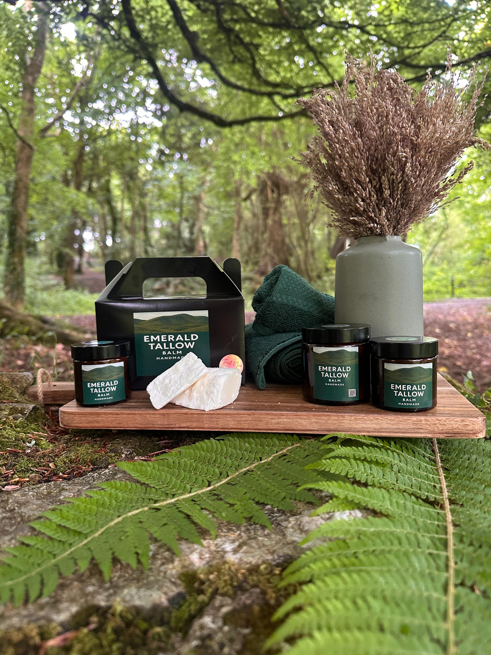 Emerald Tallow Balm product range displayed on a wooden board in a lush forest setting, featuring glass jars, gift box, raw tallow, and a ceramic vase with dried grasses.